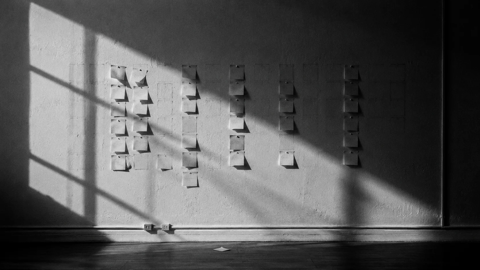 A wall of sticky notes in hard window light: some columns dense, others empty, one fallen note on the floor.