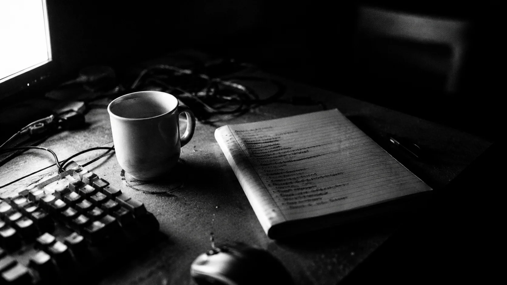 A developer's desk at night in black and white: mechanical keyboard, cold coffee, and a handwritten to-do list with most lines struck through.